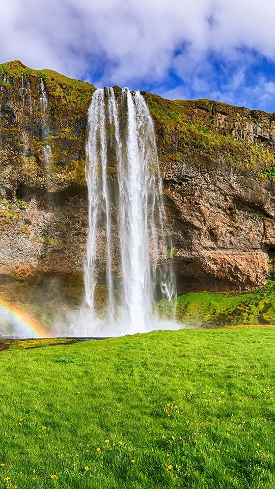 Waterfall cascading down a rocky cliff with a vibrant green grassy foreground. A rainbow arcs across the mist under a partly cloudy blue sky.