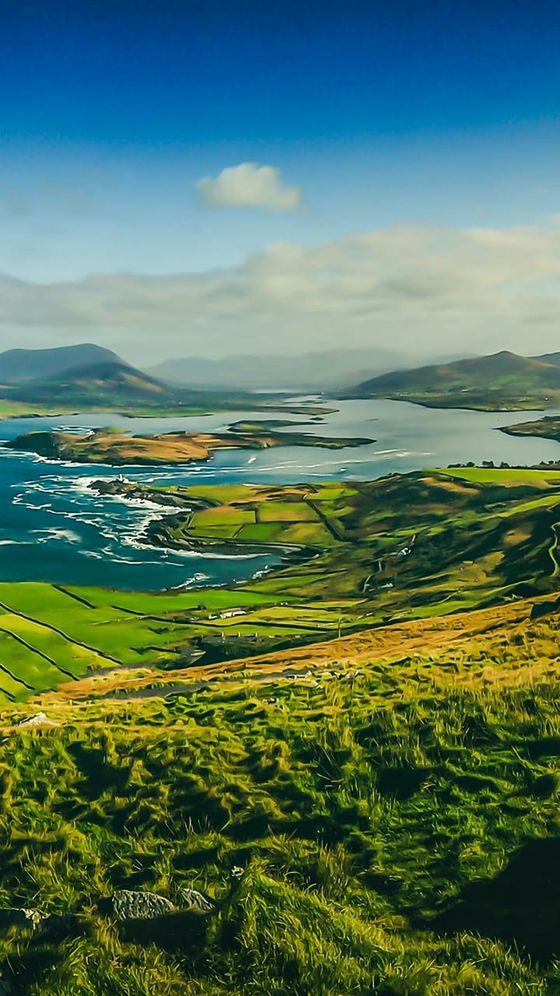 Scenic view of a coastal landscape with green fields, winding roads, and a vast ocean under a clear blue sky. Mountains are visible in the background.