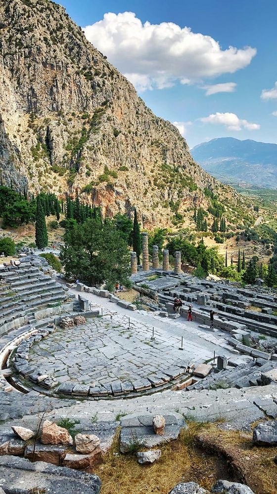 Ancient stone amphitheater with surrounding green hills, columns, and a mountainous backdrop under a partly cloudy sky.