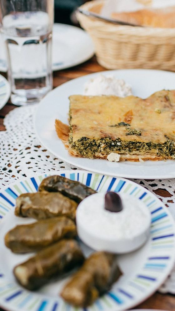 Plate with spanakopita and dip, another with stuffed grape leaves and tzatziki, on a table with a basket of bread and glasses of water.