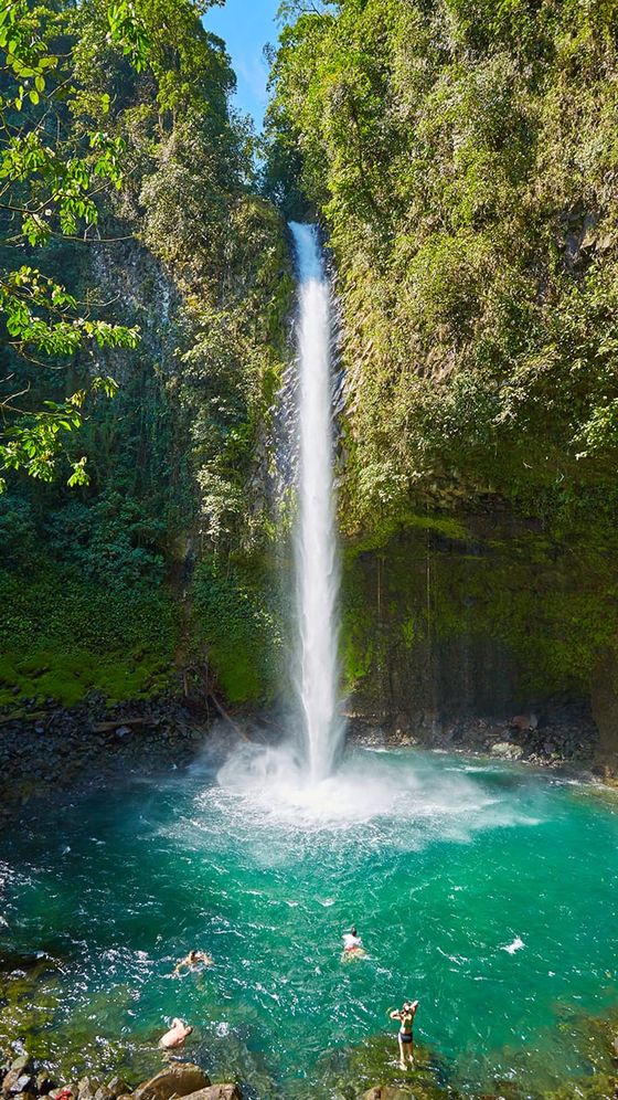 A tall waterfall cascading into a turquoise pool surrounded by lush greenery, with people swimming and enjoying the water.