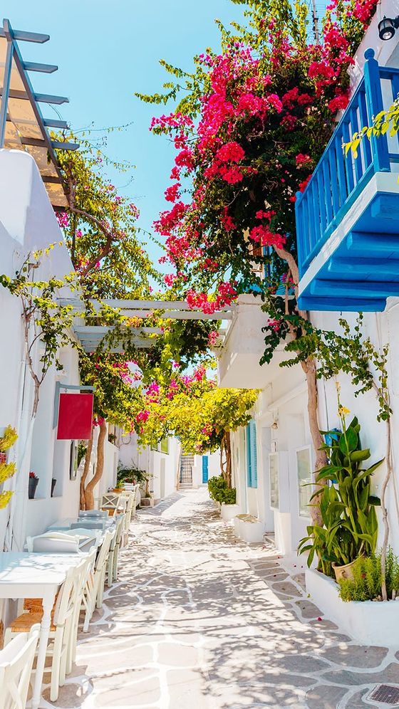 A picturesque narrow street with whitewashed buildings, vibrant bougainvillea, blue balconies, and outdoor café tables under a clear blue sky.