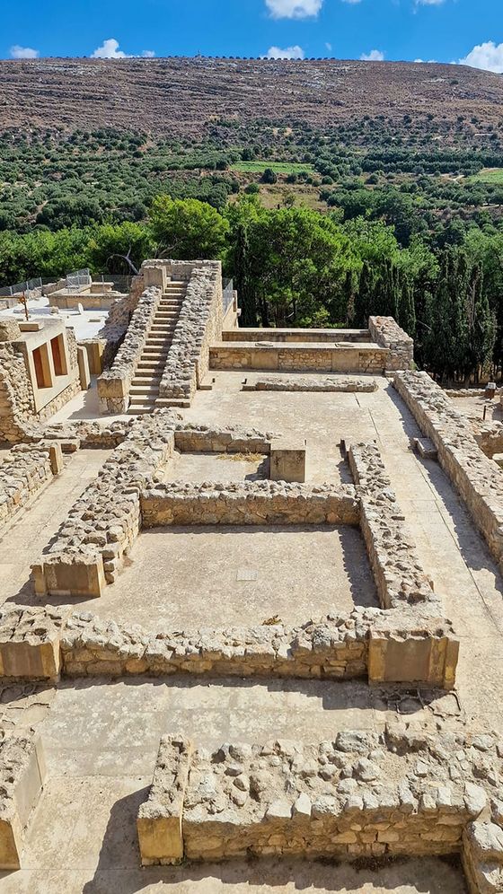 Ancient stone ruins with rectangular layout and stairs, surrounded by trees and distant hills under a blue sky.