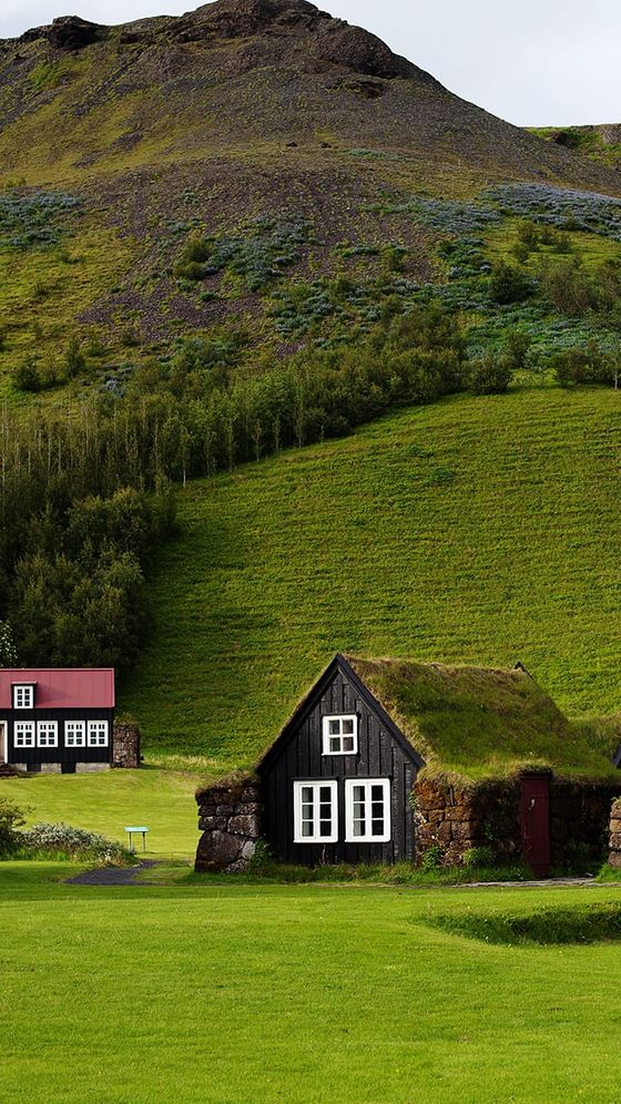 A small turf-roofed house and another with a red roof sit against a lush green hillside with a rocky hilltop in the background.