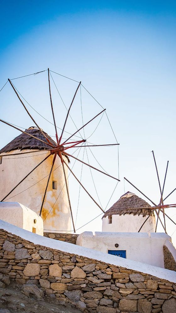 Historic white windmills with straw roofs on a stone wall in Mykonos, Greece, under a clear blue sky.