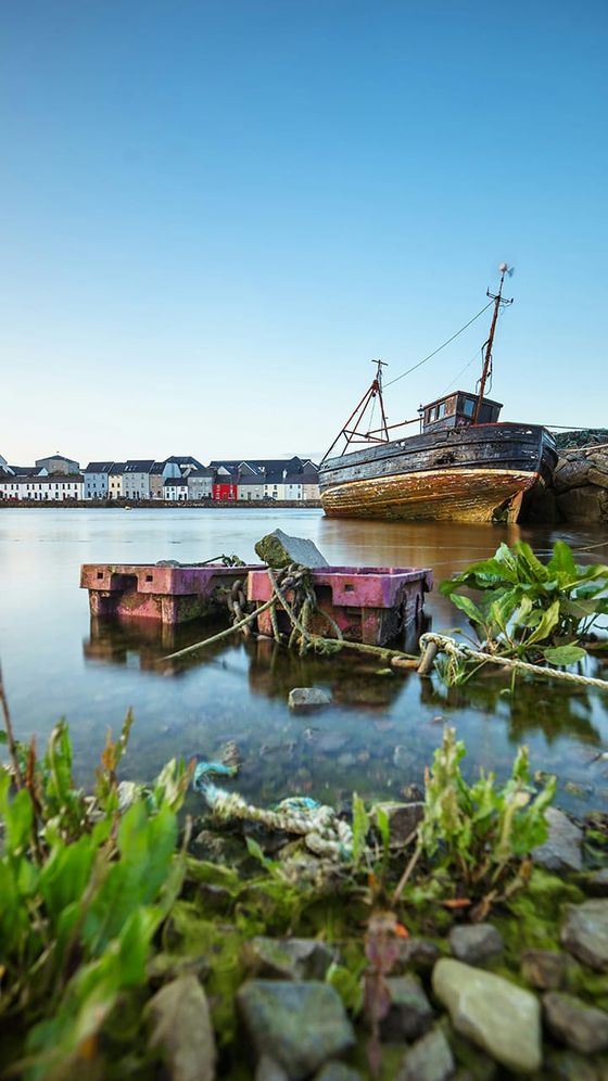 A weathered boat docked near a rocky shore with pink crates in the foreground, houses lining the background under a clear blue sky.