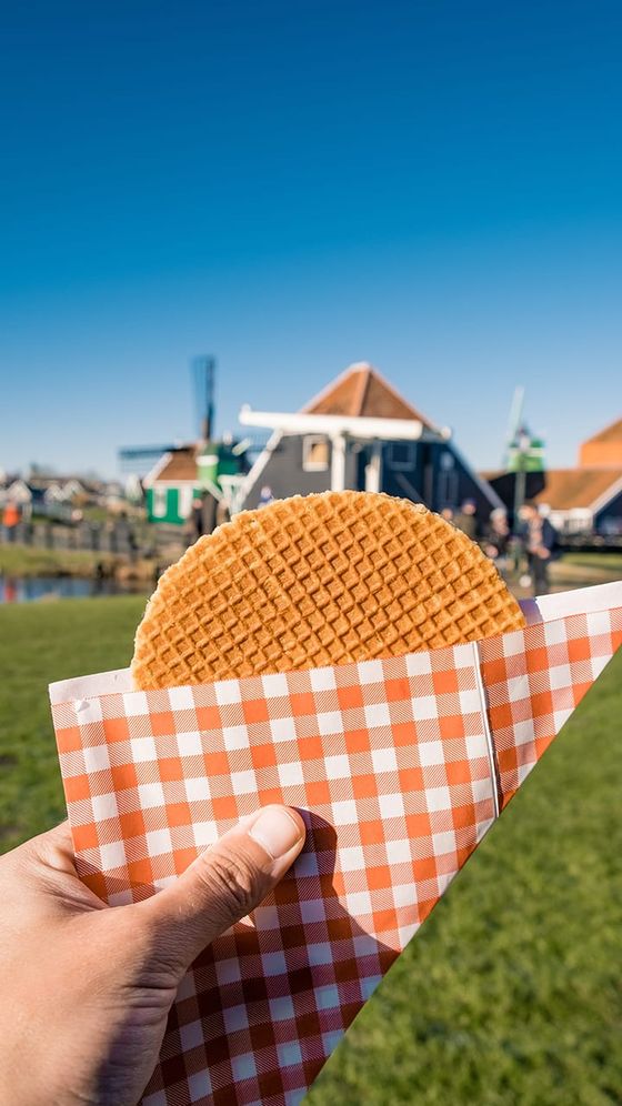 Hand holding a stroopwafel in a checkered paper wrapper, with a backdrop of traditional Dutch houses and a clear blue sky.