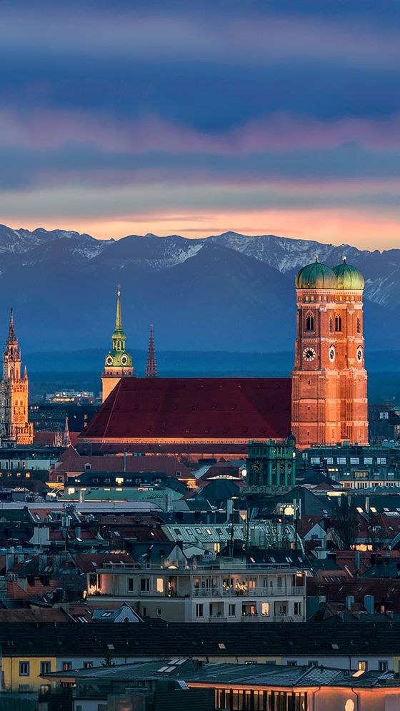 Cityscape of Munich at dusk, showcasing the Frauenkirche towers, St. Peter's Church, and Alps in the background under a vibrant sky.