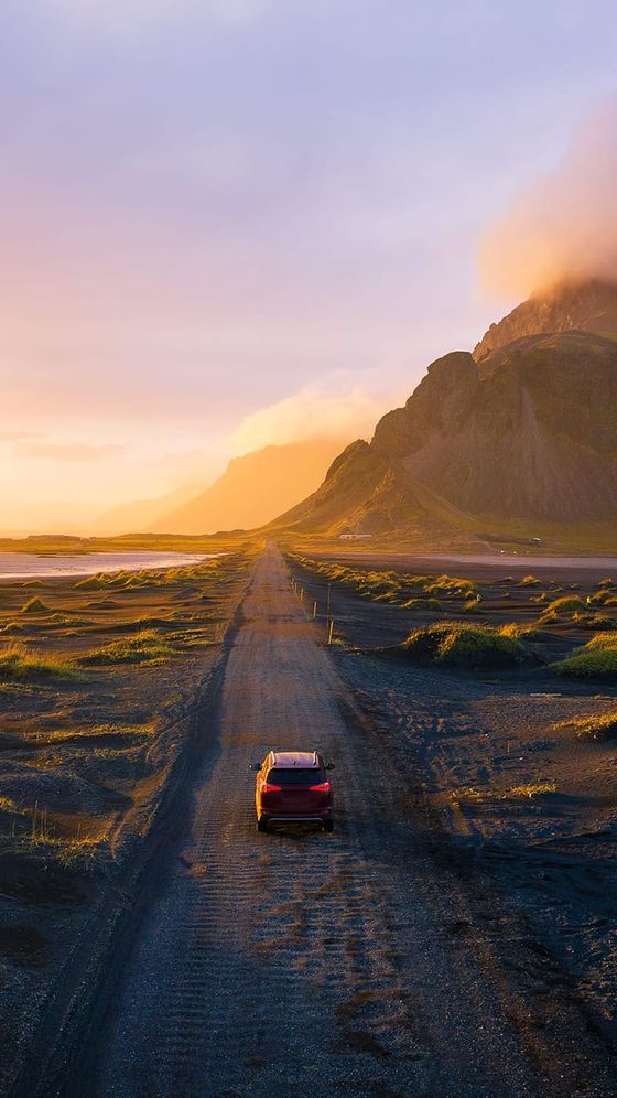 A red car travels on a gravel road through a scenic landscape with rugged mountains and a sunrise, casting a warm glow.