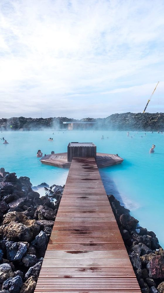 Wooden walkway leading to a small platform over a steaming geothermal lagoon, surrounded by rocky terrain. People relax in the milky blue water.