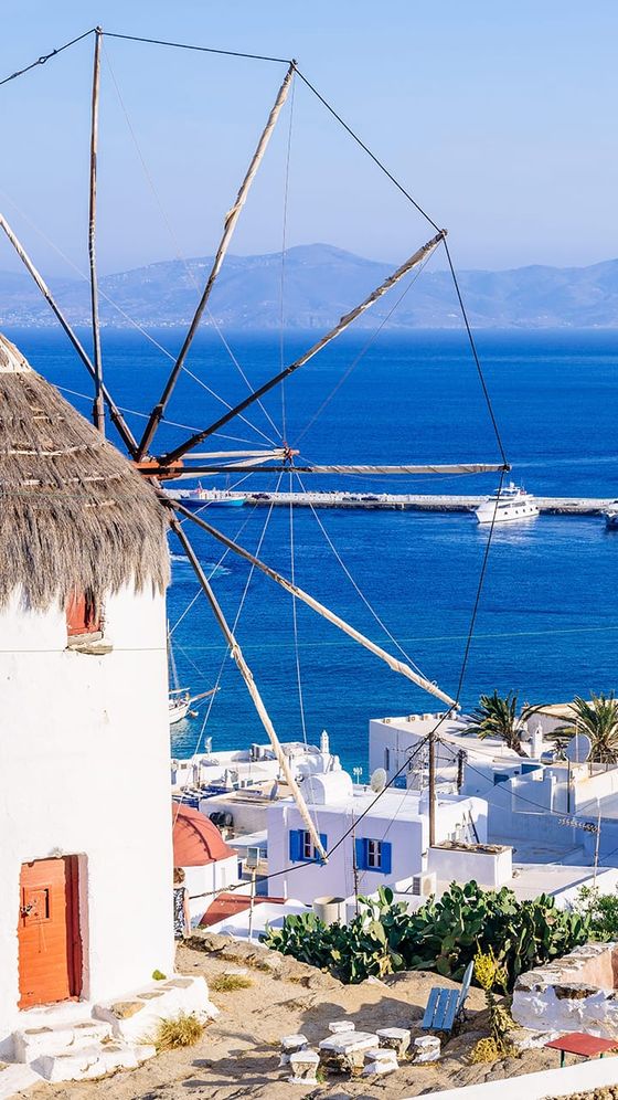 A traditional white windmill with a thatched roof overlooks the blue sea in Mykonos, Greece. White buildings and a boat are visible in the background.