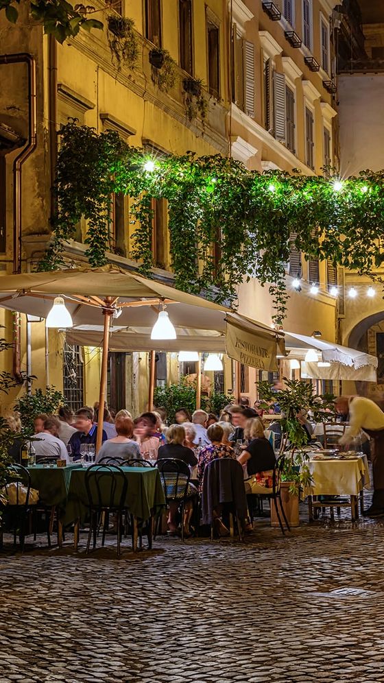 Outdoor restaurant with diners under umbrellas and string lights on a cobblestone street, with vines and greenery above. Cozy evening atmosphere.