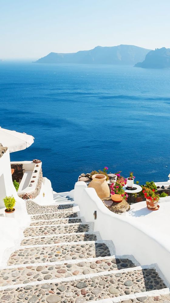 Stone steps with potted plants lead down to a white building, overlooking a vast blue sea and distant mountains under a clear sky.