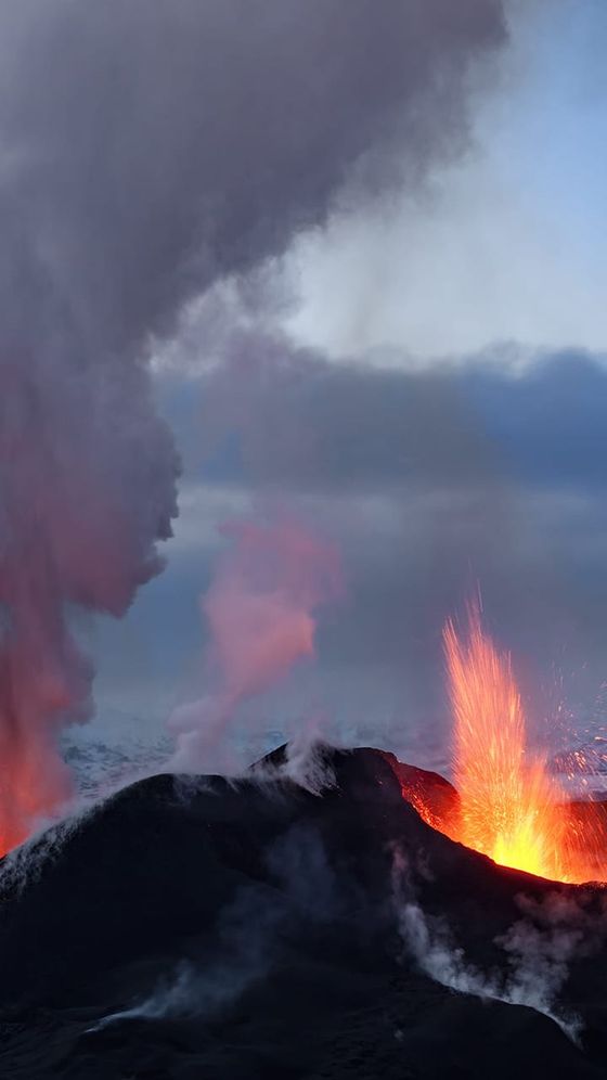 Volcano erupting with bright orange lava against dark ash clouds and a smoky sky.