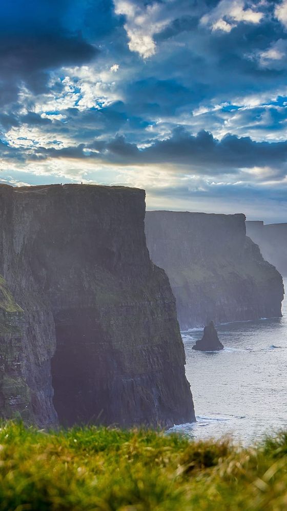 Dramatic cliffs overlooking the ocean under a cloudy sky, with green grass in the foreground and a sea stack visible in the water.