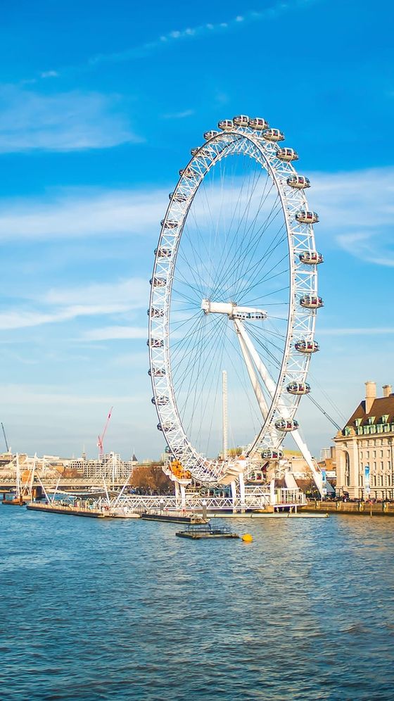 Large Ferris wheel beside a river with a blue sky backdrop, surrounded by buildings and cranes.