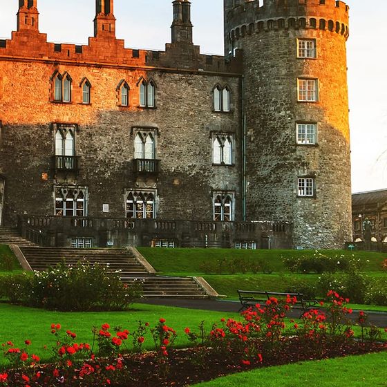 Historic castle with tall stone tower, surrounded by lush gardens and red flowers, under a clear blue sky at sunset.