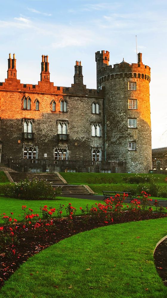 Historic castle with tall stone tower, surrounded by lush gardens and red flowers, under a clear blue sky at sunset.