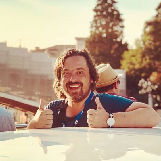 Man smiling with thumbs up, wearing a blue shirt and watch, outside with trees and another person in a hat in the background.
