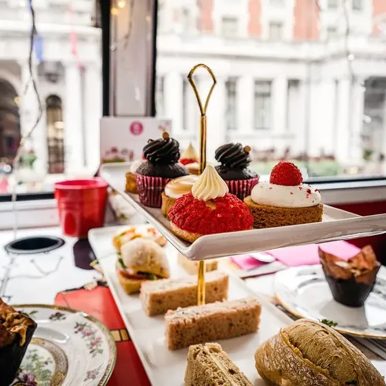 A tiered tray with assorted desserts and sandwiches on a table in a cafe, with a view of a building through the window in the background.