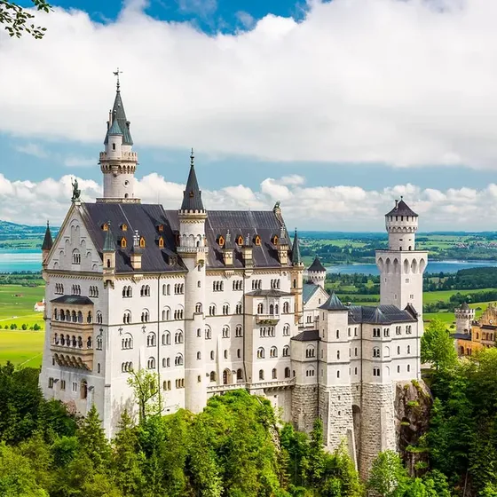 A grand castle with tall spires set on a lush hillside, overlooking a landscape of green fields and a distant lake under a cloudy sky.
