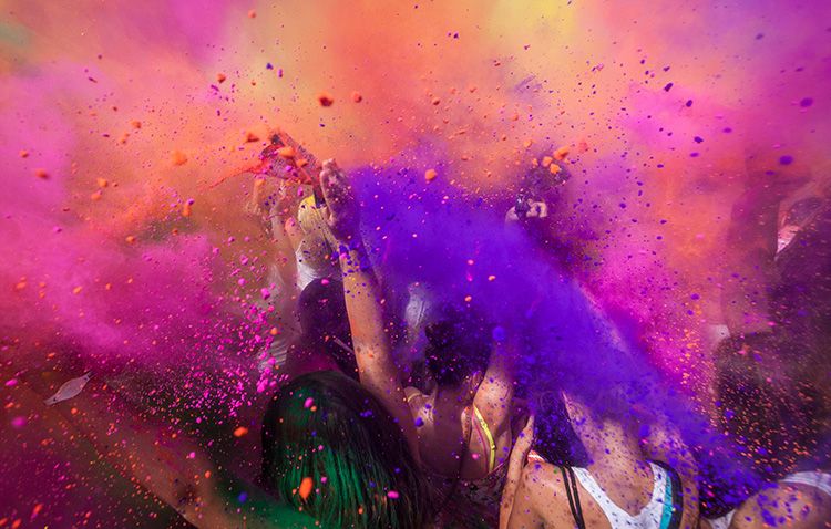 People celebrating Holi, throwing vibrant colored powders in the air, creating a lively explosion of purple, pink, orange, and yellow hues.