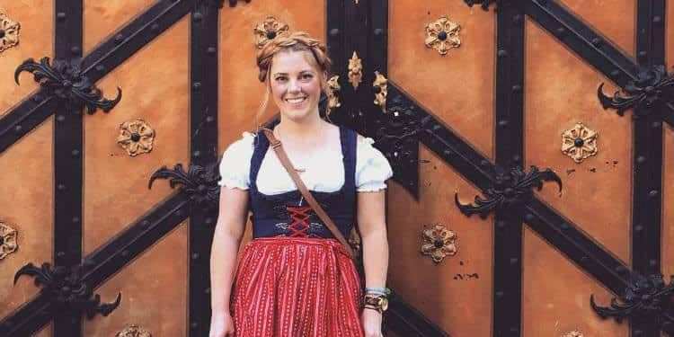Woman in traditional Bavarian dress stands smiling in front of an ornate wooden door with decorative ironwork.