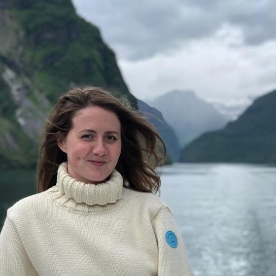 A woman in a white sweater smiles on a boat with a scenic view of cloudy skies, mountains, and a tranquil body of water in the background.