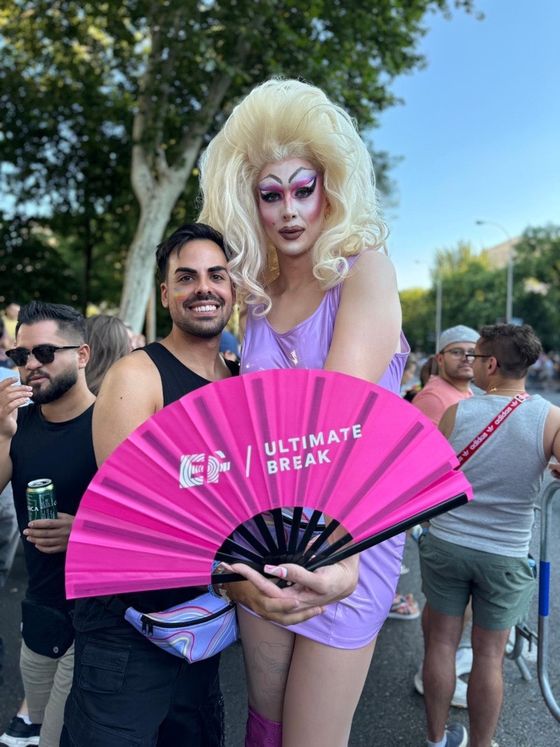 A person in drag with elaborate makeup and a blonde wig holds a large pink fan reading "Ultimate Break," posing with a smiling attendee outdoors.