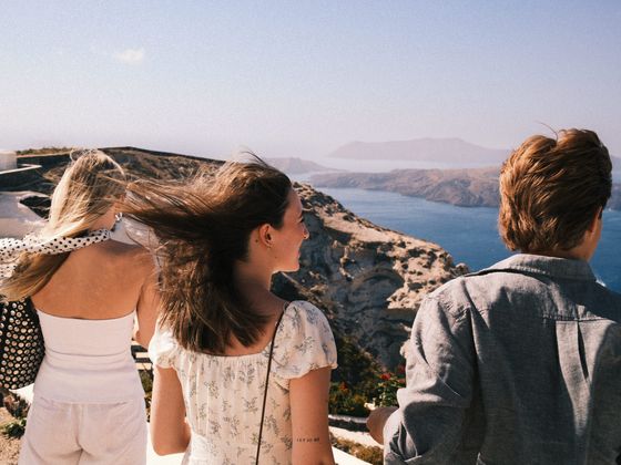 Three people enjoy a scenic view of cliffs and the sea on a sunny day, with the wind blowing through their hair.