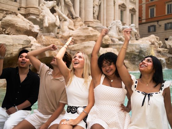 Five people joyfully sitting by a fountain, throwing coins into the water. The background features intricate stone sculptures.