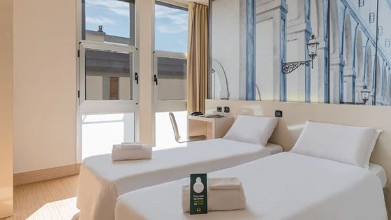 Bright hotel room with two neatly made single beds, a desk with a chair, a large window, and a mural of an architectural design on the wall.