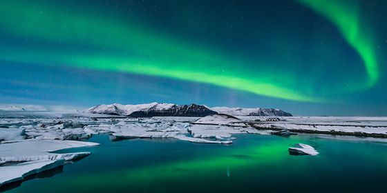 A vibrant green aurora borealis illuminates the night sky over a snowy landscape and icy water, with distant mountains in the background.