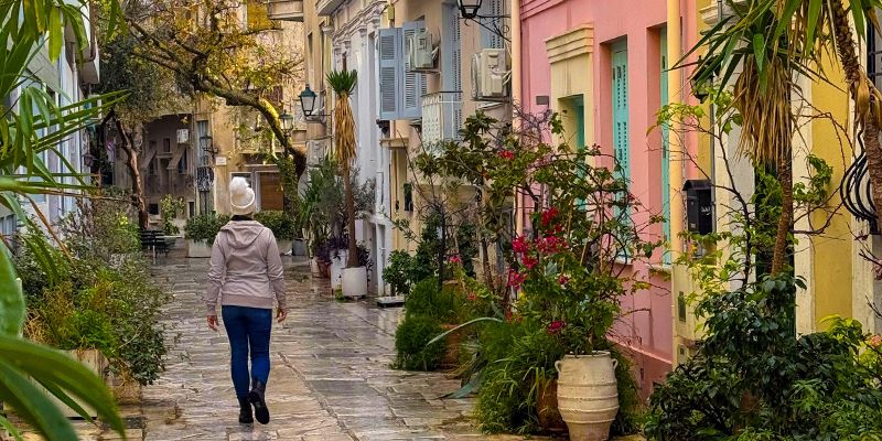 Woman in a hat walks down a charming, narrow street lined with colorful buildings and lush greenery.