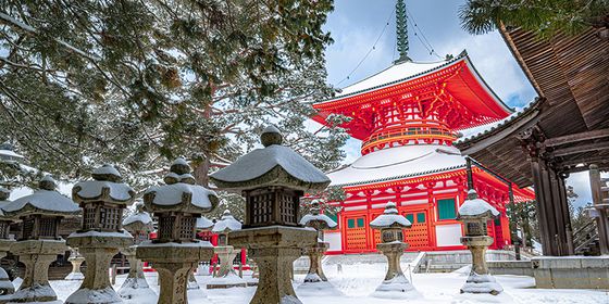 Snow-covered stone lanterns lead to a vibrant red pagoda with a tiered roof in a serene, snowy landscape.
