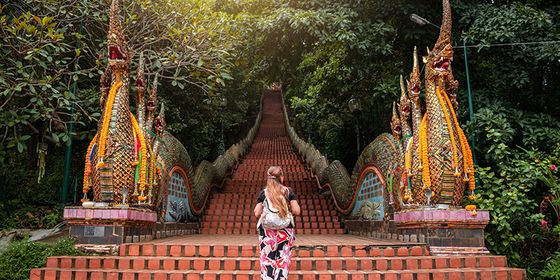 A woman in a floral dress climbs a brick staircase flanked by ornate dragon sculptures, surrounded by lush greenery.