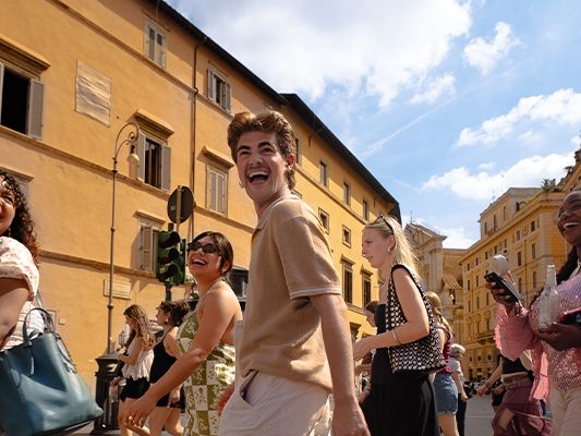 A group of joyful people walking on a sunny street with historic yellow buildings in the background.