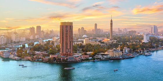 Aerial view of a city skyline with a river winding through, featuring tall buildings, boats on the water, and a colorful sunset sky.