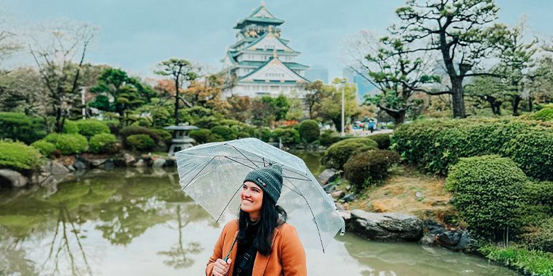 A woman holding a transparent umbrella stands by a pond, with a traditional Japanese castle in the background amidst lush greenery.