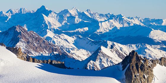 Snow-covered mountain range under a clear blue sky, showcasing rugged peaks and deep valleys in bright sunlight.