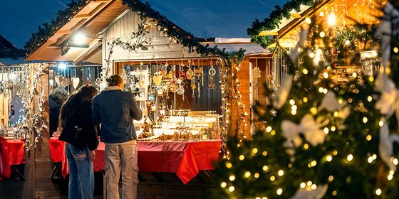People browsing illuminated stalls at a festive outdoor market, with holiday decorations and a lit Christmas tree in the foreground.