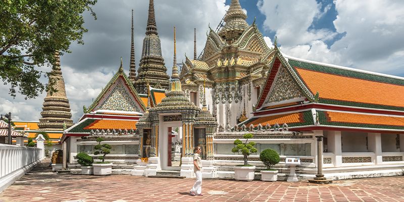 A person walks near ornate temples with colorful roofs and spires at Wat Pho in Bangkok, Thailand, under a partly cloudy sky.