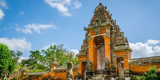 Ancient temple gate with intricate carvings, surrounded by lush greenery, under a bright blue sky with scattered clouds.