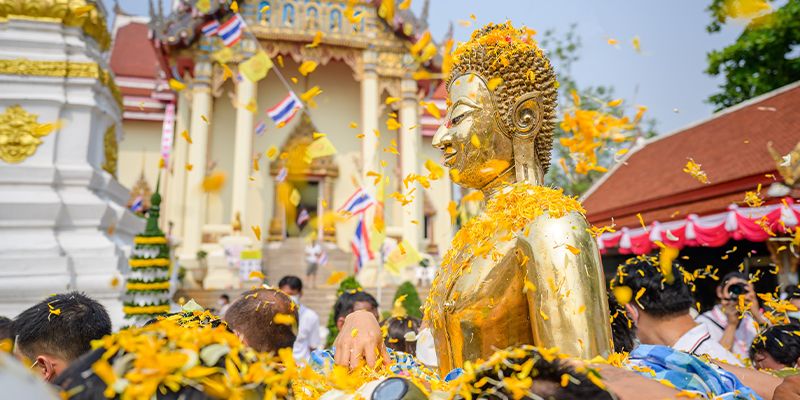 Golden Buddha statue adorned with yellow flower petals, surrounded by people during a festive celebration at a temple, with Thai flags visible.