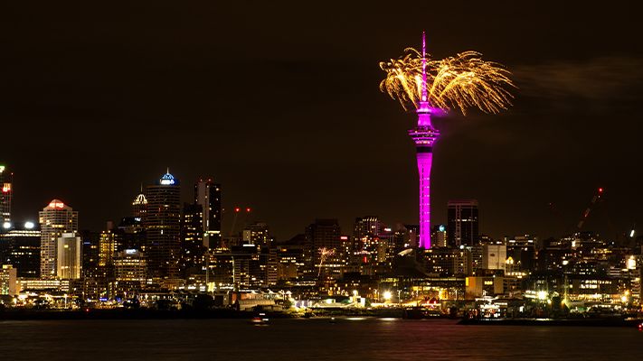 A vibrant city skyline at night with a tall tower lit in purple, fireworks bursting at its top against the dark sky.