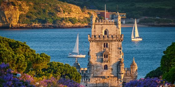 Belem Tower in Lisbon, with sailboats on the Tagus River and lush greenery in the foreground under a clear blue sky.