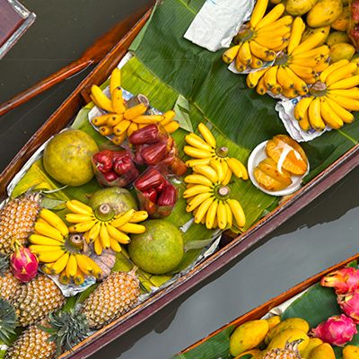 Aerial view of boats at a floating market with vibrant tropical fruits, including bananas, pineapples, and coconuts. A vendor in a straw hat is visible.