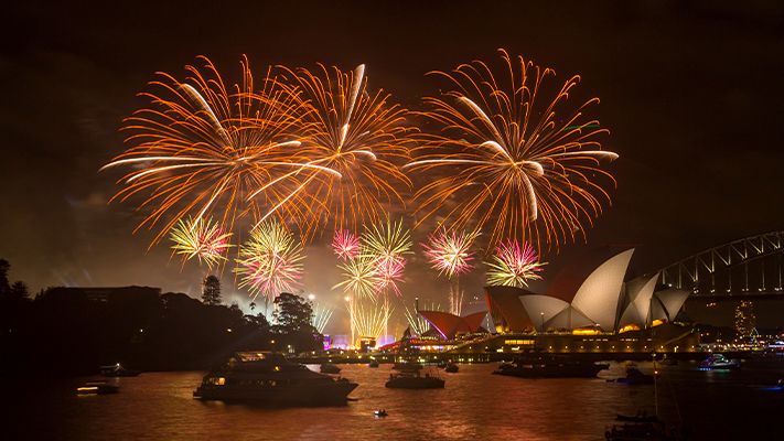 Colorful fireworks light up the night sky over Sydney Harbour with the Opera House and Harbour Bridge in view, reflecting on the water.