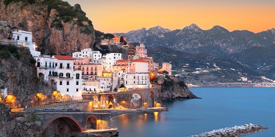 Scenic view of a coastal town at sunset, with colorful buildings perched on cliffs overlooking calm blue waters and distant mountains.
