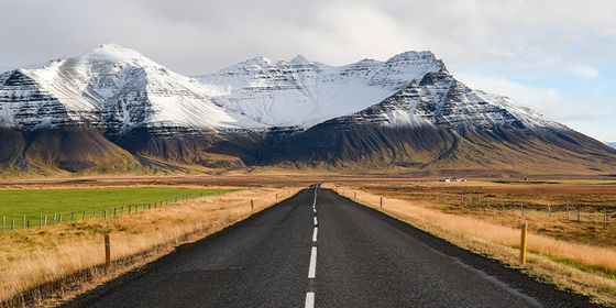 A straight road leads towards snow-capped mountains under a partly cloudy sky, flanked by fields of grass and fences.
